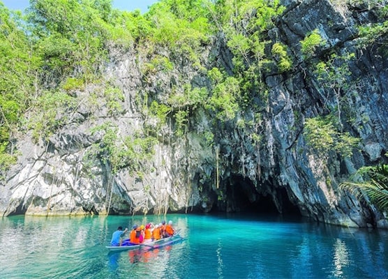 View of Puerto Princesa Subterranean River in Palawan, showcasing dark waters flowing through a dramatic limestone cave, surrounded by dense greenery and towering cliffs, with visitors exploring the natural wonder