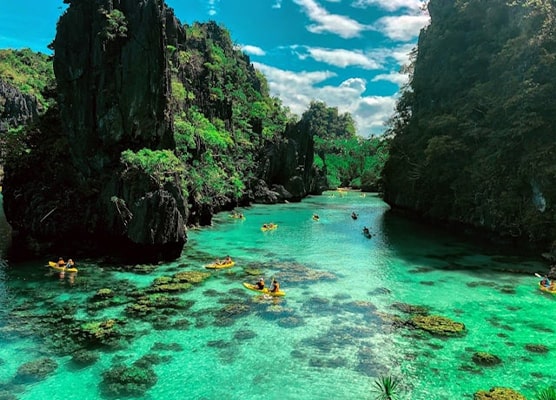 View of Big Lagoon in El Nido, Palawan, featuring turquoise waters, towering limestone cliffs, and tourists kayaking under a clear blue sky.